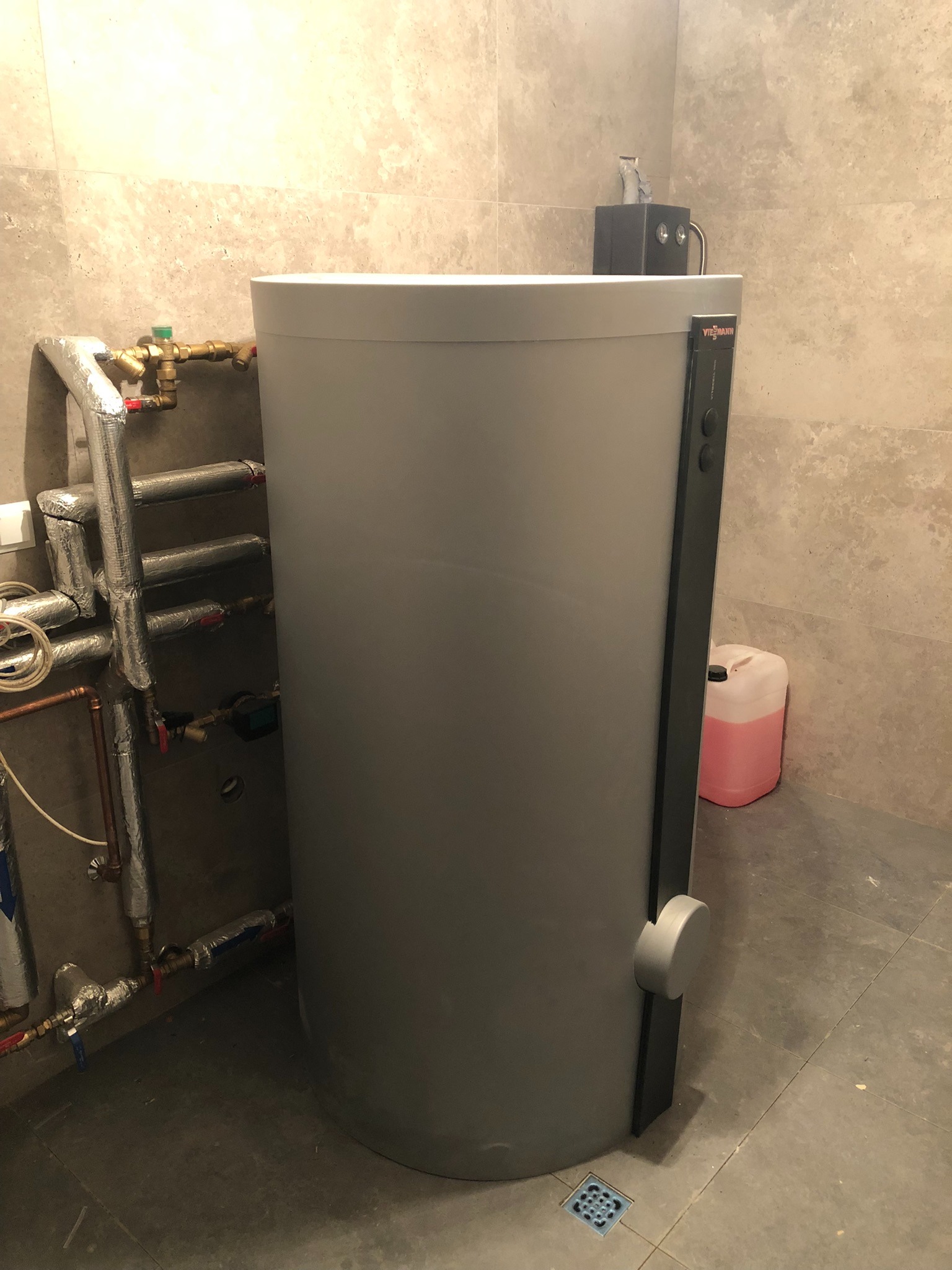 Szary zasobnik ciepłej wody Viessmann z widocznymi rurami instalacji grzewczej w pomieszczeniu wyłożonym płytkami. U dołu kratka ściekowa.