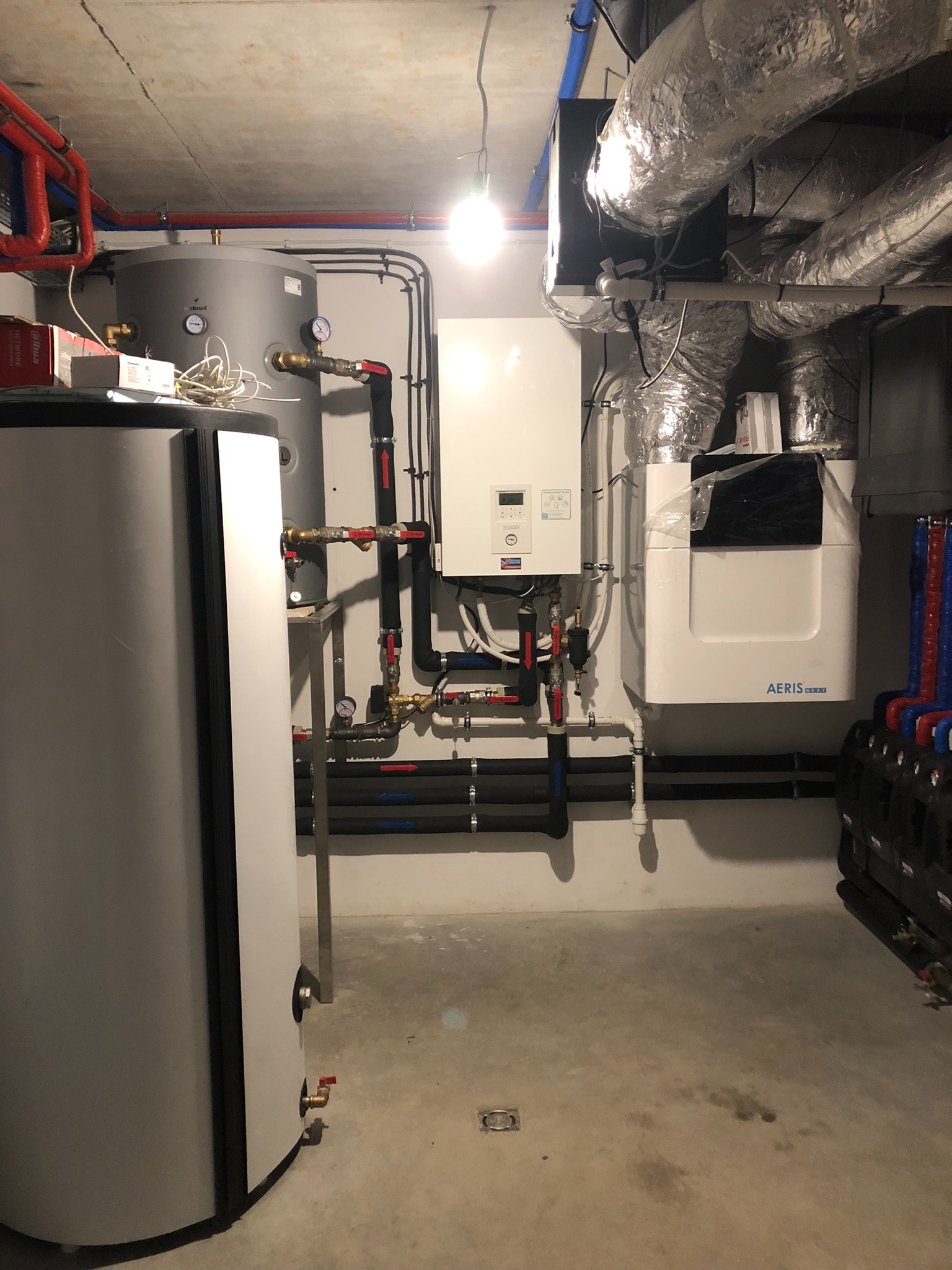 Zestaw urządzeń: zbiornik buforowy, pompa ciepła Aeris i instalacja grzewcza z rurami w izolacji, w kotłowni z betonową posadzką i wentylacją.