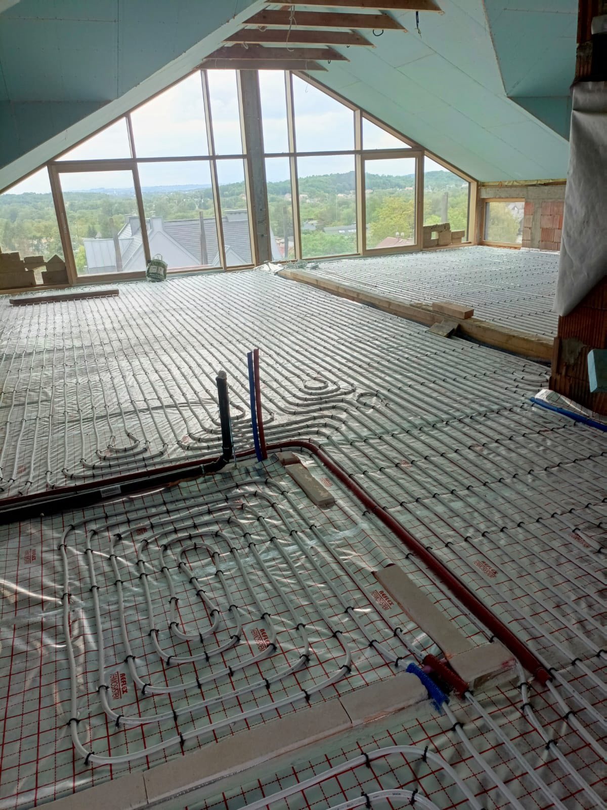 Instalacja ogrzewania podłogowego w jasnym pomieszczeniu z dużym oknem. Białe rurki ułożone na folii z czerwoną siatką, widoczne elementy instalacji hydraulicznej.