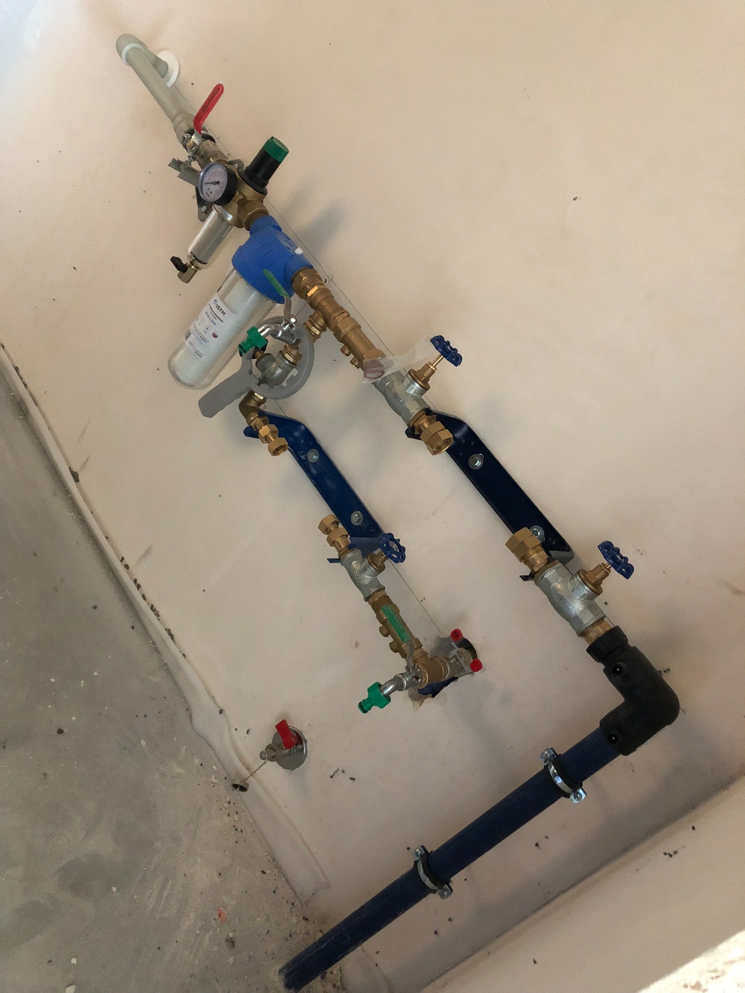 Nowoczesna instalacja wodna z filtrem, manometrem i zaworami, zamontowana na ścianie w surowym wnętrzu. Widoczne elementy miedziane, stalowe i plastikowe.