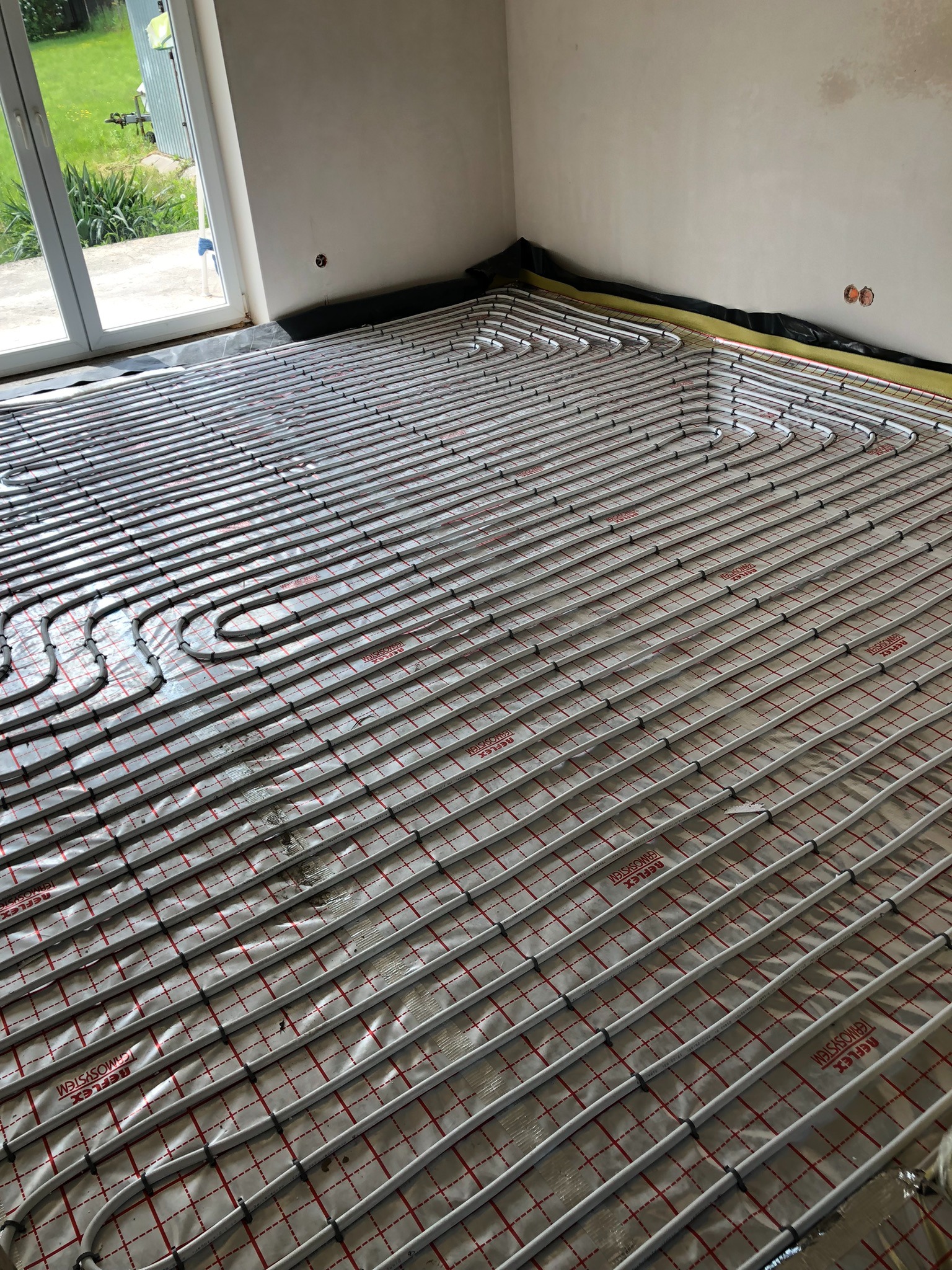 Ułożone rury ogrzewania podłogowego na folii refleksyjnej w nowym budynku, widoczne okno i przygotowane punkty elektryczne na ścianie.