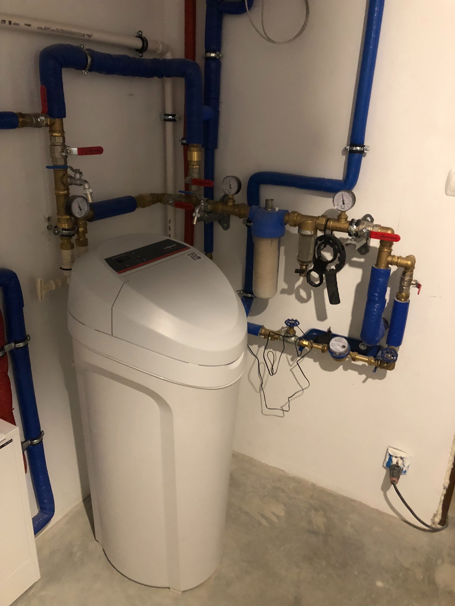 Biały zmiękczacz wody Viessmann z niebieskimi rurami i manometrami na ścianie w pomieszczeniu. Widoczne złącza, zawory i filtr. Szare podłoże, gniazdko elektryczne.