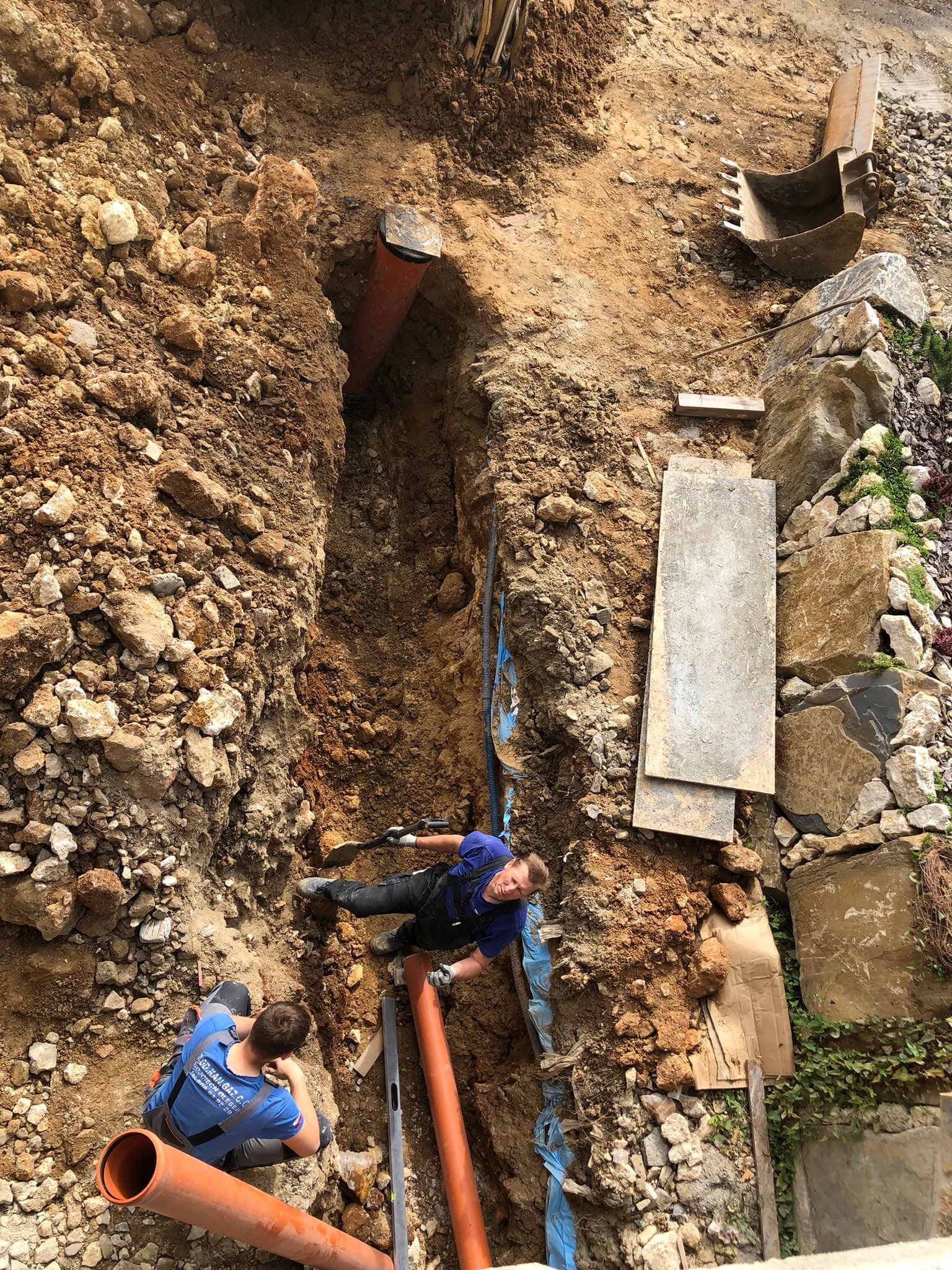 Montaż rur w wykopie ziemnym z użyciem poziomicy. Widoczni dwaj pracownicy w trakcie pracy. W tle fragment koparki i kamienne elementy dekoracyjne.