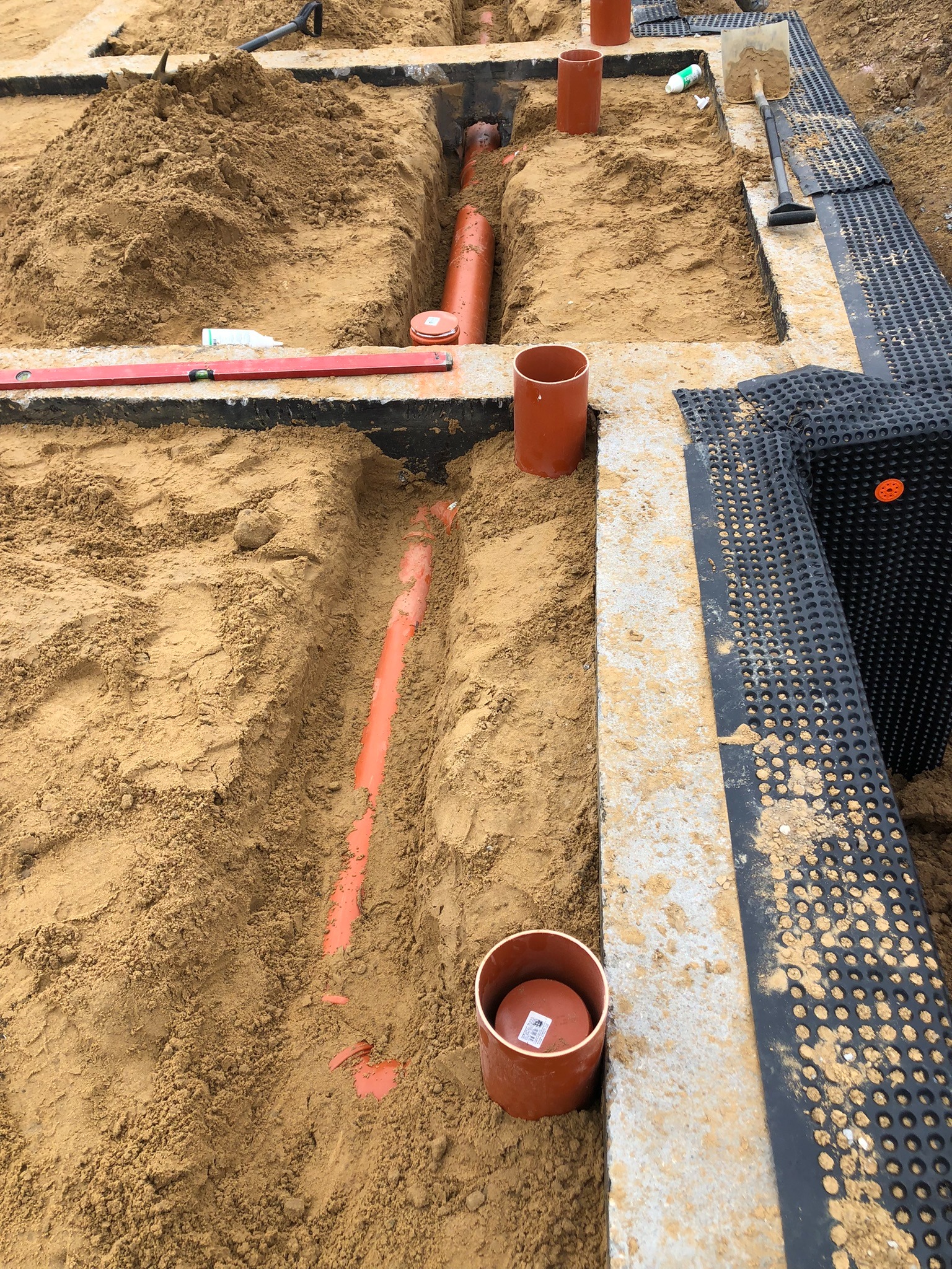 Pomarańczowe rury kanalizacyjne ułożone w wykopie z piasku, widoczne narzędzia i czarna folia izolacyjna. Etap prac instalacyjnych.