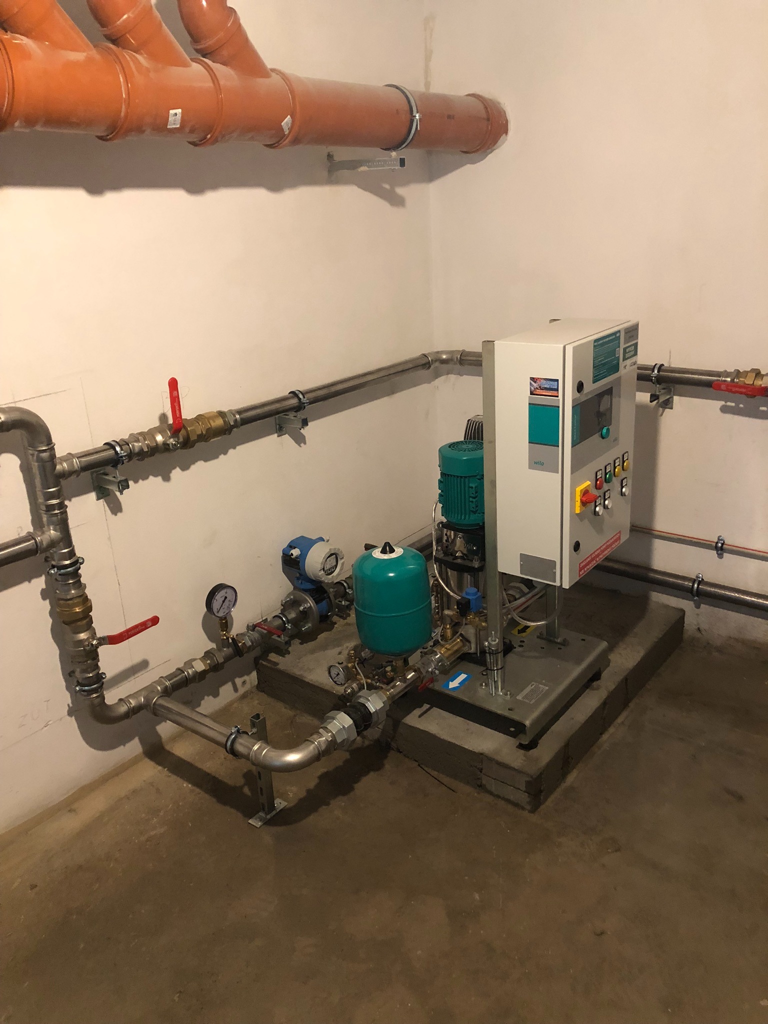 Złożona instalacja wodociągowa z rurami miedzianymi i plastikowymi, zaworami, manometrami oraz pompą Wilo na betonowym fundamencie w pomieszczeniu technicznym.