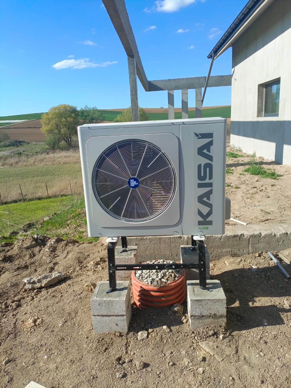 Kompaktowa pompa ciepła Kaisai, zamontowana na betonowych blokach w Myślenicach. Widoczne elementy instalacji i krajobraz w tle. Nowoczesne ogrzewanie.
