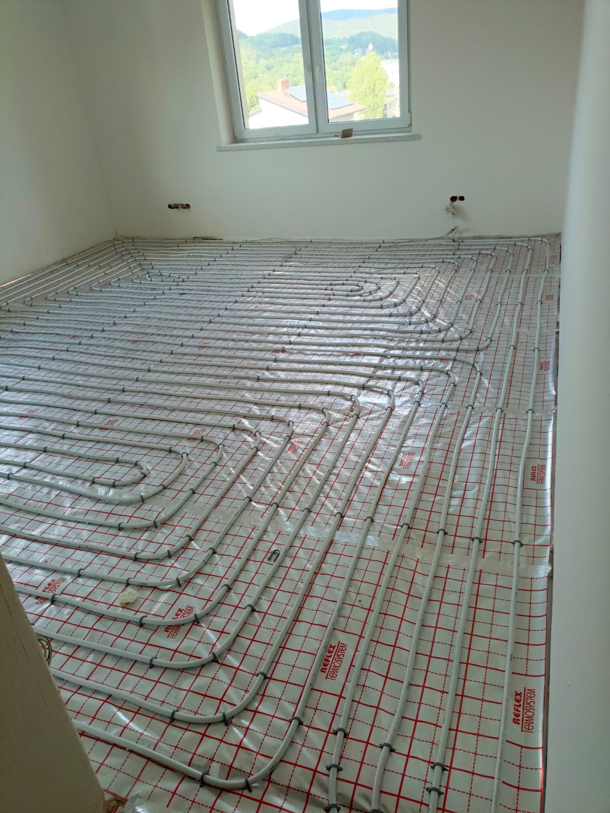 Białe rury ogrzewania podłogowego ułożone na folii z czerwoną kratką w jasnym pokoju z oknem i widokiem na zalesione wzgórza w tle. Etap instalacji przed wylewką.