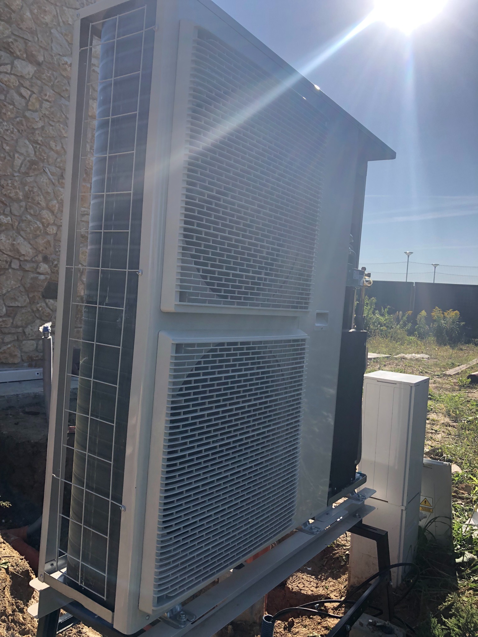 Zewnętrzna jednostka pompy ciepła, zamontowana na metalowej konstrukcji przy budynku z kamienia, w słoneczny dzień. Widoczne elementy instalacji i skrzynka elektryczna.
