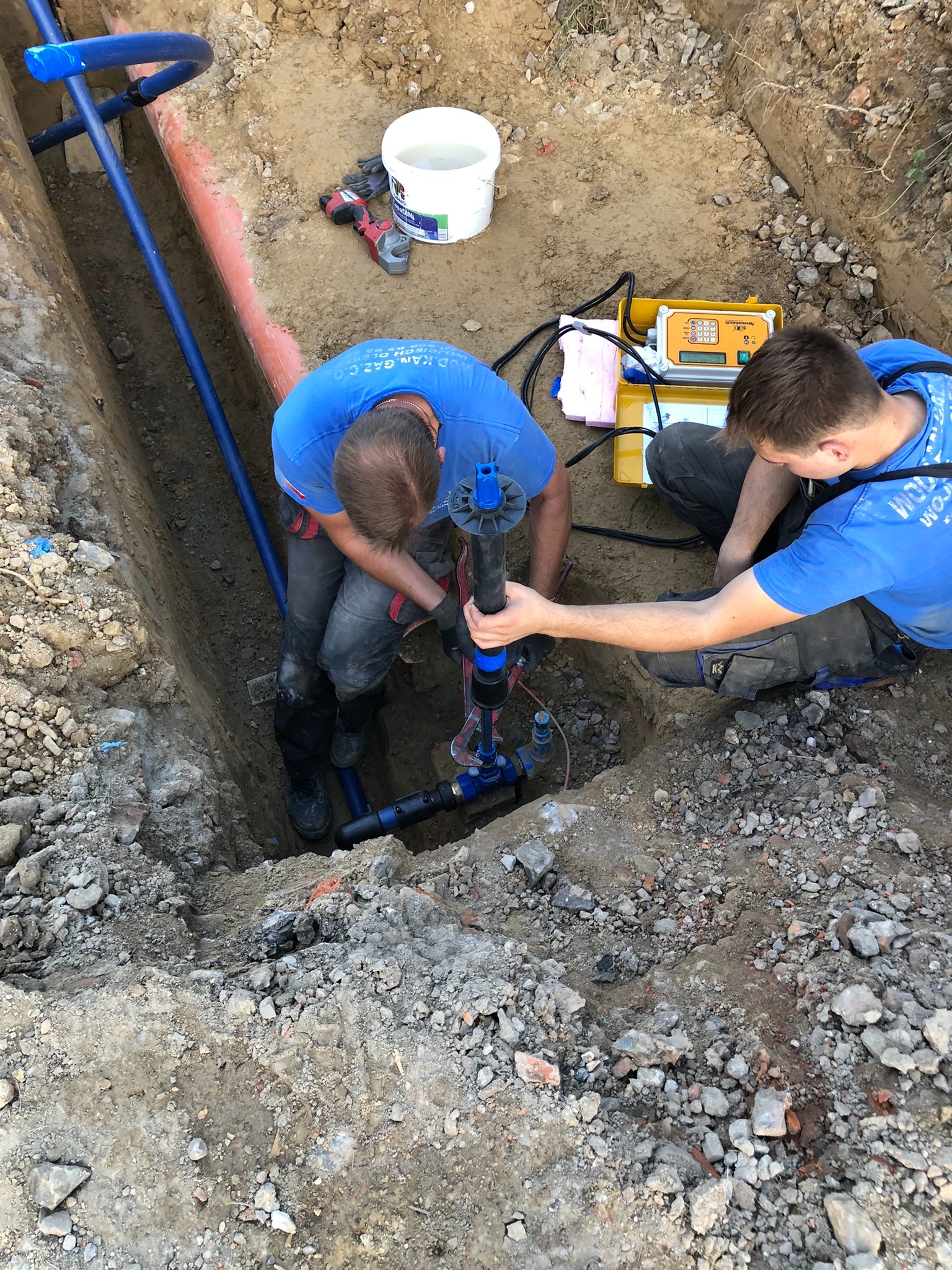 Dwóch instalatorów w niebieskich koszulkach łączy rury w wykopie, używając specjalistycznego narzędzia. Obok leży urządzenie pomiarowe w żółtej obudowie.