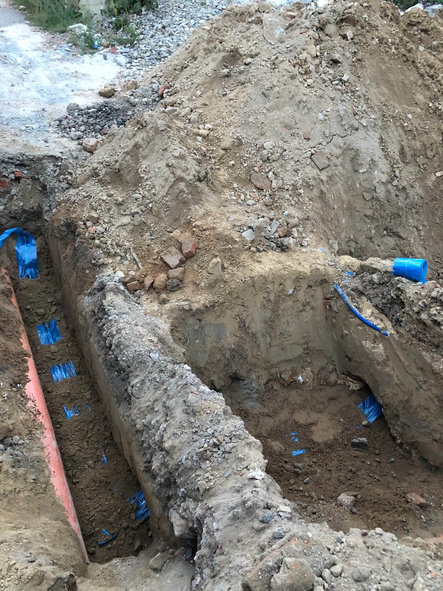 Wykop z widoczną pomarańczową rurą oraz niebieskimi zabezpieczeniami izolacyjnymi, ziemia z gruzem i kamieniami.
