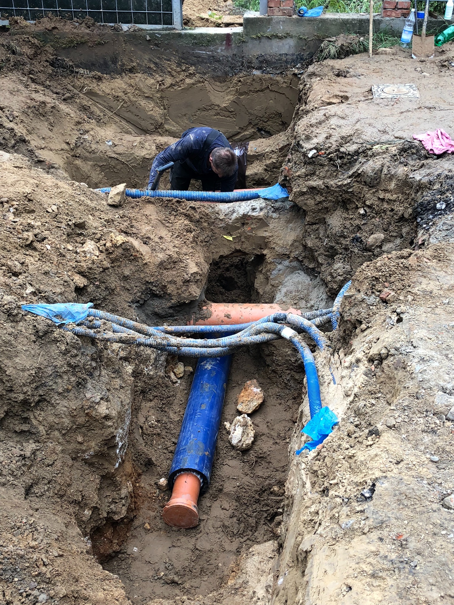 Głęboki wykop w ziemi z ułożonymi rurami instalacyjnymi w kolorze niebieskim i pomarańczowym, pracownik w czarnym kombinezonie pochylony nad instalacją.