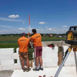 Dwóch pracowników w pomarańczowych koszulkach z logo firmy MURDOR w Gliwicach, wyrównuje ścianę z białych bloczków za pomocą poziomicy laserowej i łaty murarskiej na tle zielonych pól i błękitnego...