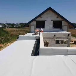 Prace na dachu płaskim domu w trakcie budowy: dwóch mężczyzn w krótkich spodenkach układa białą membranę dachową na podłożu z czarnej papy, w tle widać niezadaszoną konstrukcję budynku z bloczków...