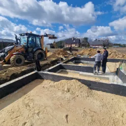 Budowa fundamentów domu jednorodzinnego: widoczny wykop, zbrojenie, betonowe bloczki pomalowane na czarno, piasek, oraz koparko-ładowarka CASE w tle, dwóch robotników w trakcie pracy.