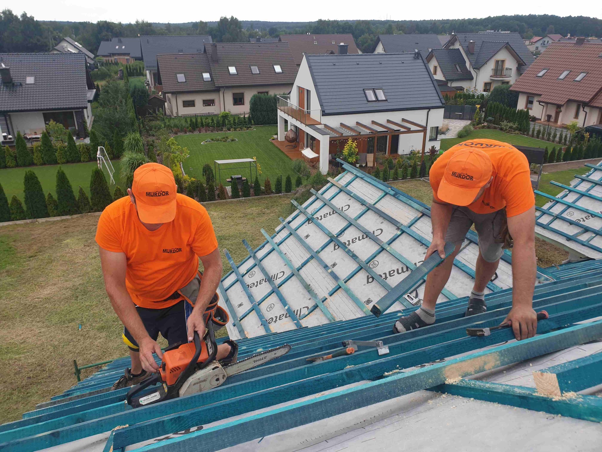 Dwóch pracowników w pomarańczowych koszulkach i czapkach z logo firmy montuje konstrukcję drewnianą dachu, używając piły spalinowej i młotka, na tle domów jednorodzinnych.