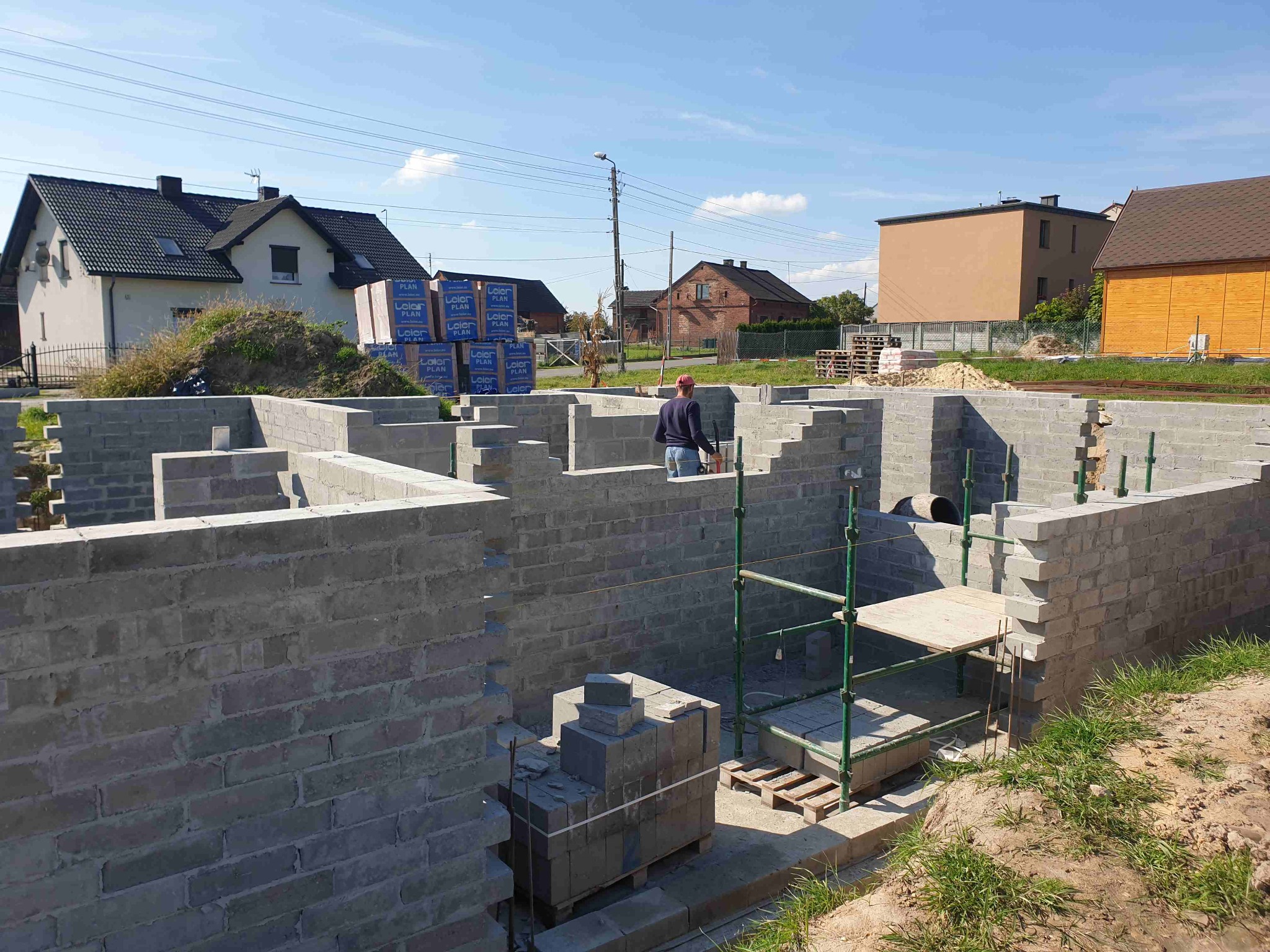 Budowa fundamentów z bloczków betonowych na działce z widokiem na okoliczne domy, widoczne rusztowanie i palety z materiałem.