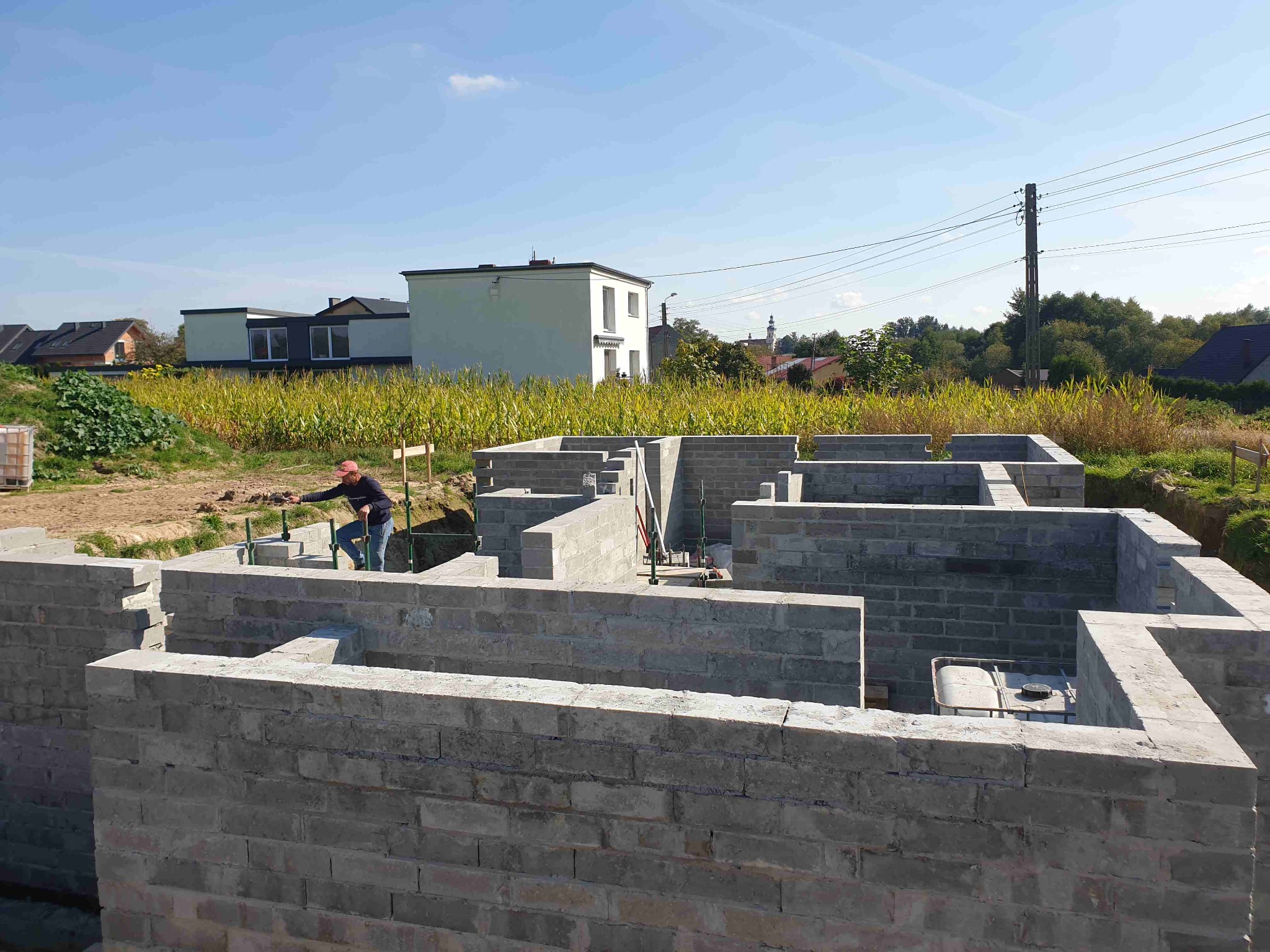 Wznoszenie fundamentów domu z bloczków betonowych, widoczny pracownik w czerwonej czapce i narzędzia, w tle zabudowa jednorodzinna i pole kukurydzy.