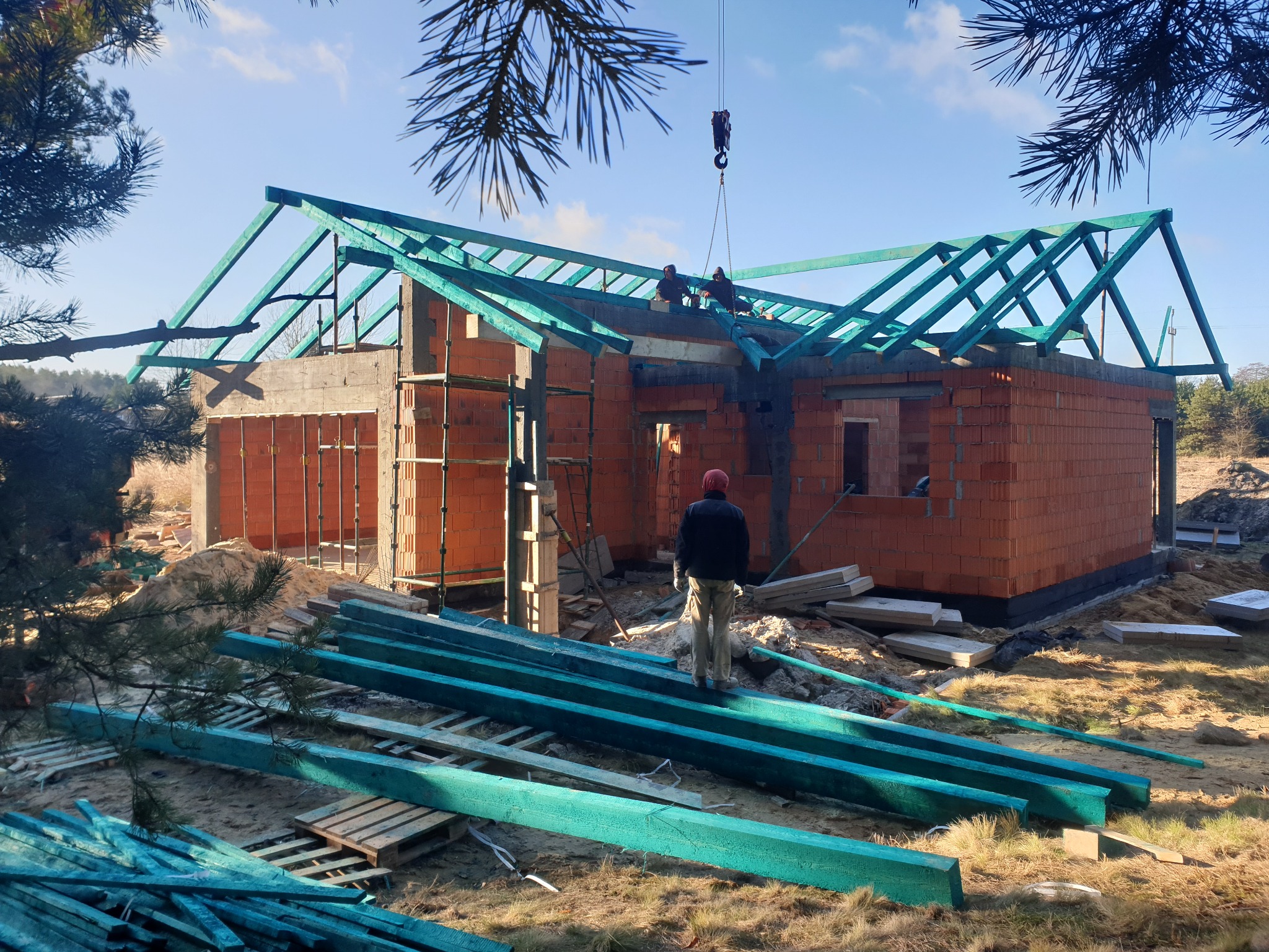 Wznoszenie konstrukcji dachu na budynku z czerwonej cegły, widoczny dźwig podnoszący elementy, na pierwszym planie pomalowane na zielono elementy konstrukcyjne.