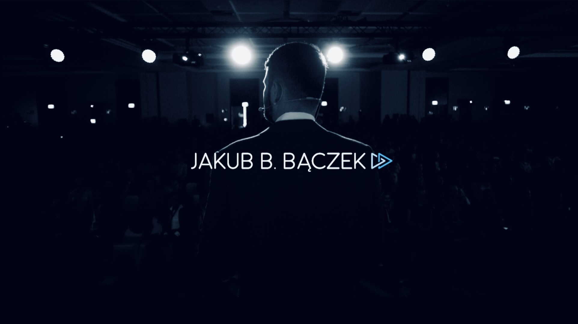 Prelegent Jakub B. Bączek na scenie, widok z tyłu, mikrofon nagłowny, publiczność w tle, oświetlenie sceniczne.