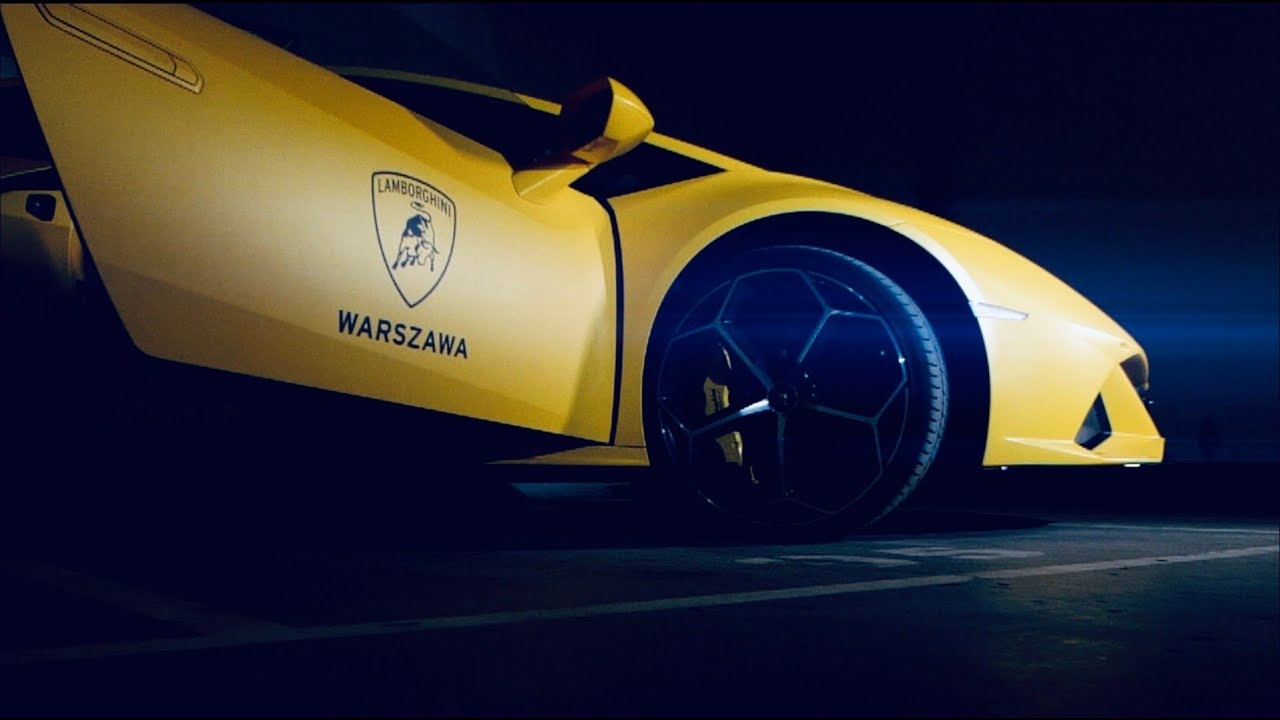 Żółte Lamborghini Huracan z logo i napisem Warszawa na drzwiach, widok z boku w nocy, oświetlone reflektorem.