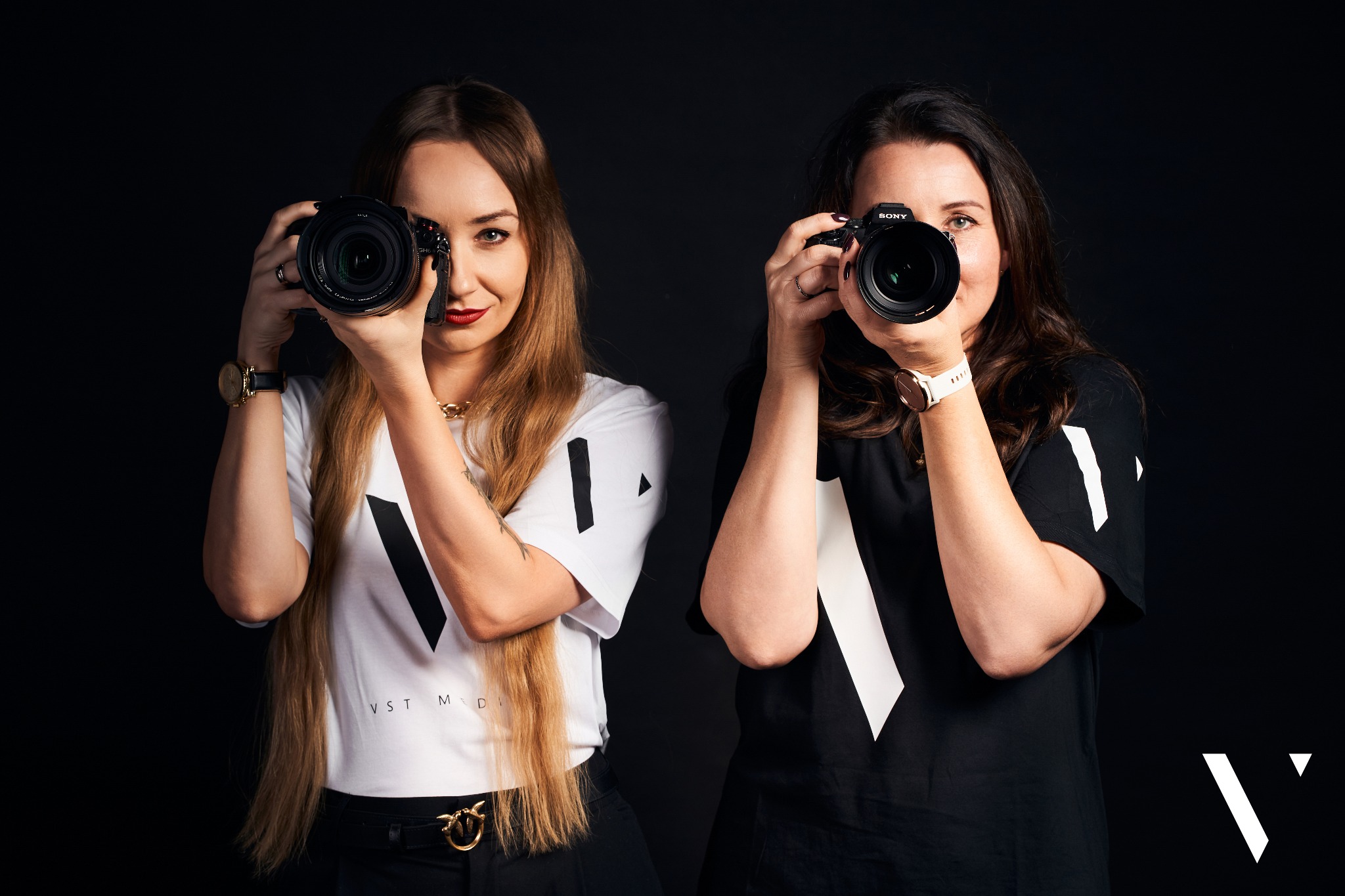 Dwie kobiety, blondynka z czerwonymi ustami i brunetka, trzymają aparaty Sony z obiektywami skierowanymi w stronę widza na czarnym tle. Ubrane są w koszulki z logo firmy VST MEDIA.