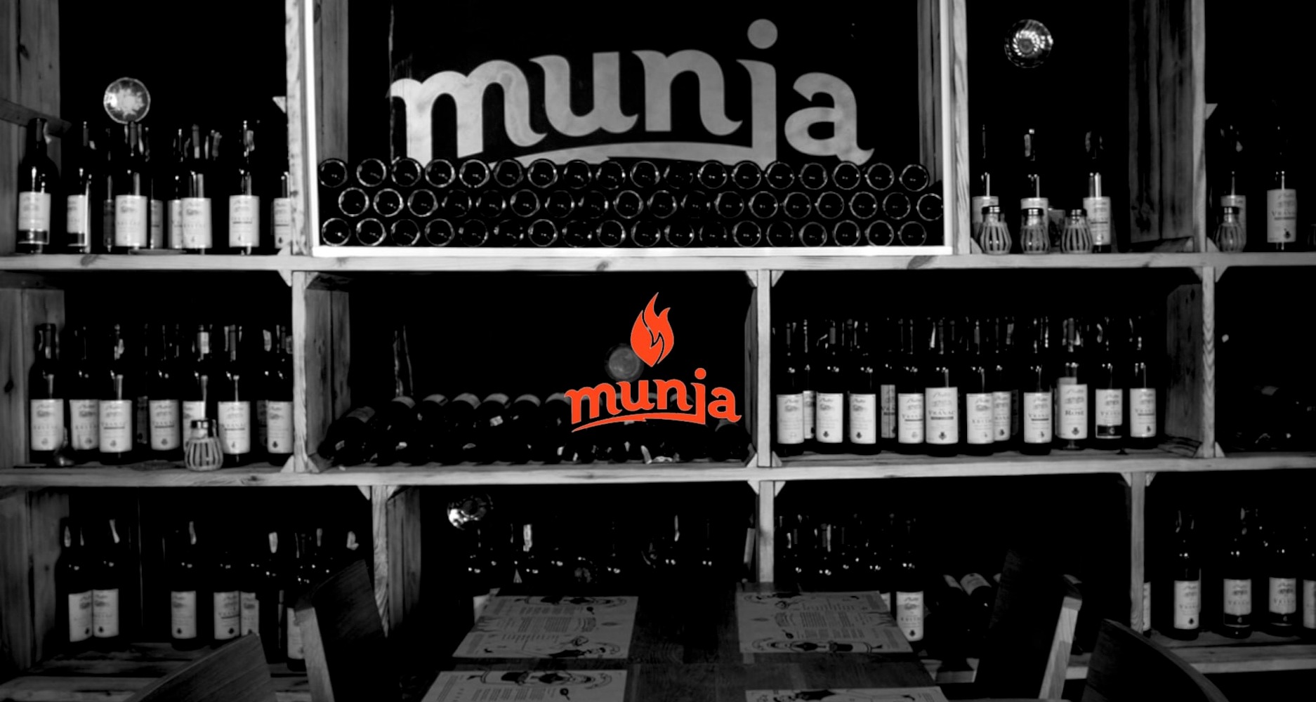 Czarno-białe zdjęcie wnętrza restauracji z drewnianymi półkami pełnymi butelek wina, nad półkami widoczne podświetlone logo 'munja'.