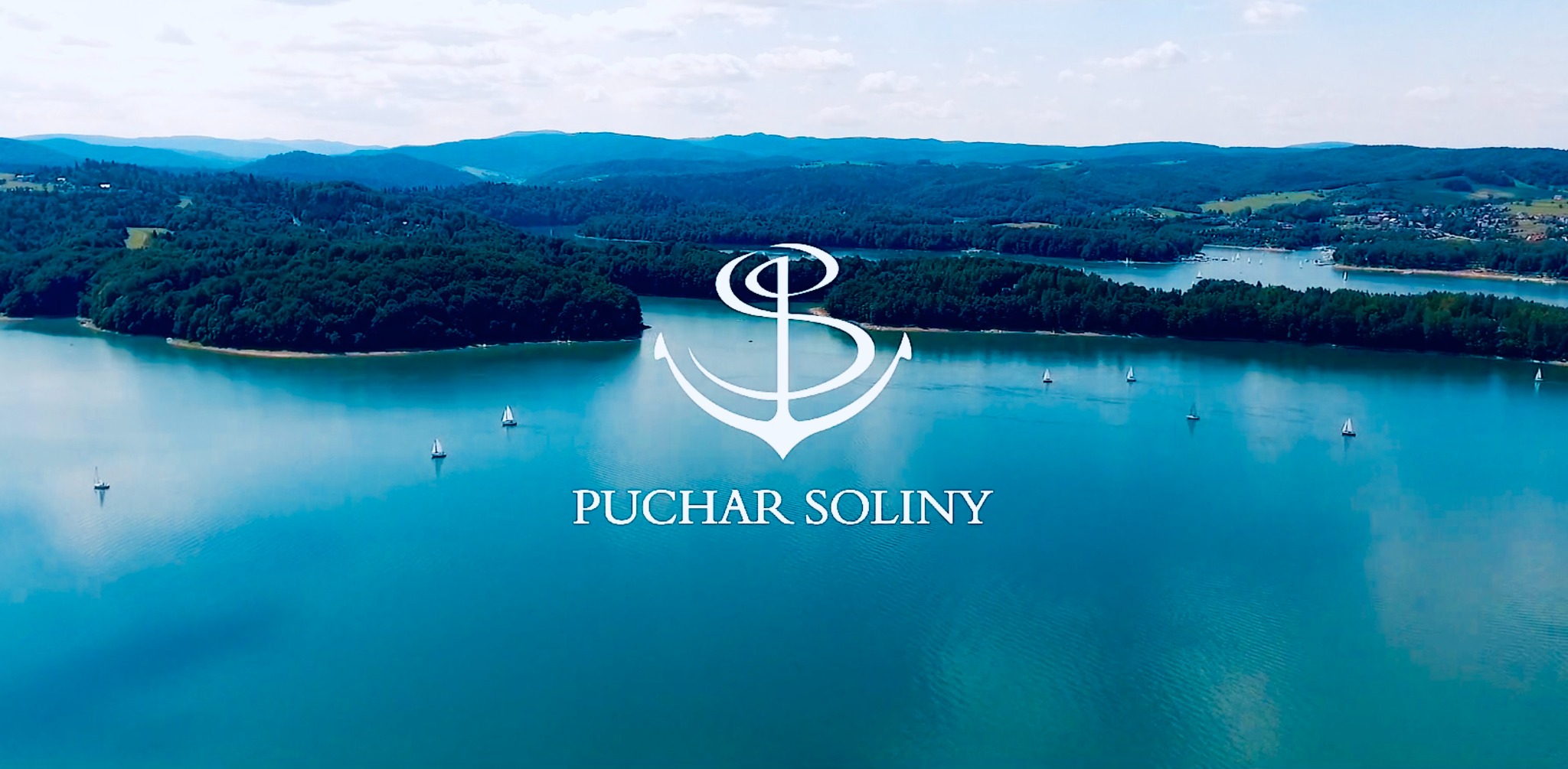 Szeroki plan jeziora Solińskiego z żaglówkami, otoczonego zalesionymi wzgórzami, z nałożonym logo 'Puchar Soliny' z symbolem kotwicy.