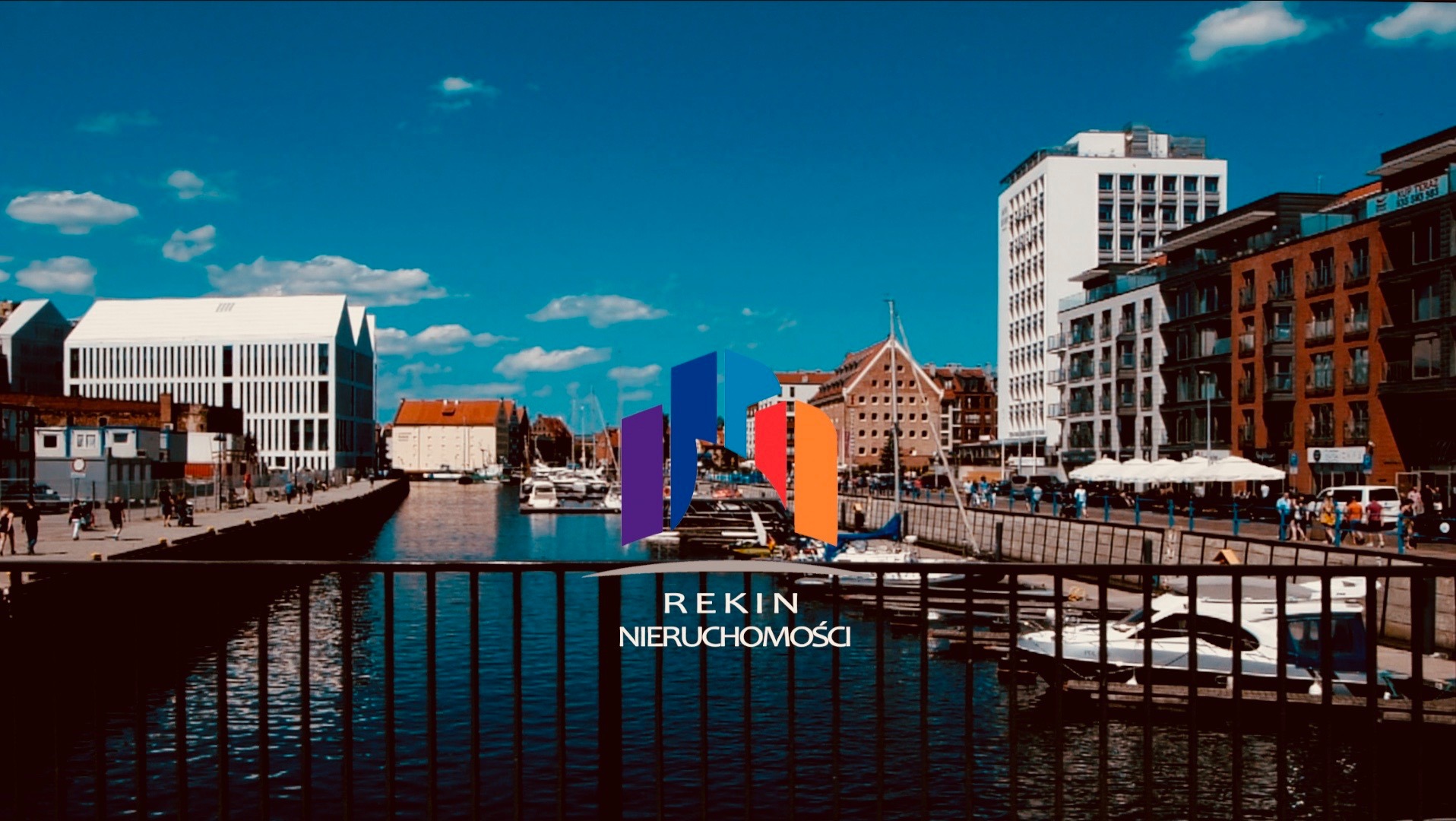 Panorama miasta z kanałem wodnym, budynkami o różnej architekturze i logo firmy REKIN Nieruchomości na pierwszym planie, widoczne zza metalowej barierki.