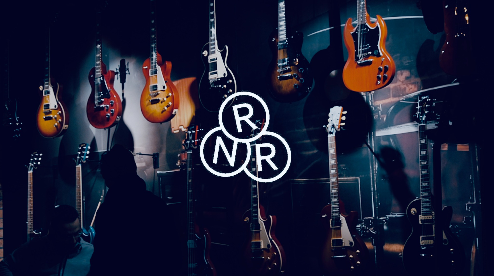 Wystawa gitar elektrycznych różnych marek i kolorów, oświetlona z przodu, z logo 'RNR' na pierwszym planie, sylwetki osób w tle.