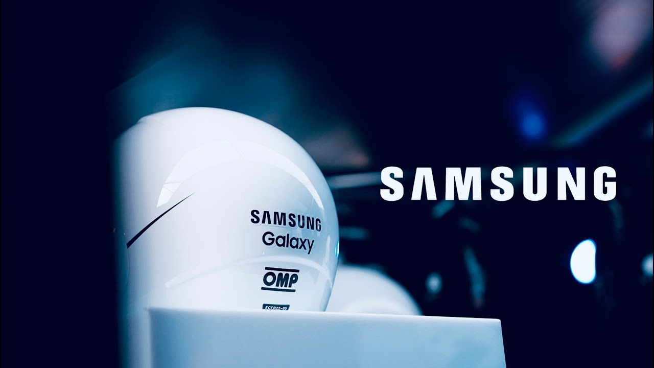 Biały kask wyścigowy Samsung Galaxy OMP na białym postumencie, z dużym napisem SAMSUNG w tle, oświetlony niebieskim światłem.