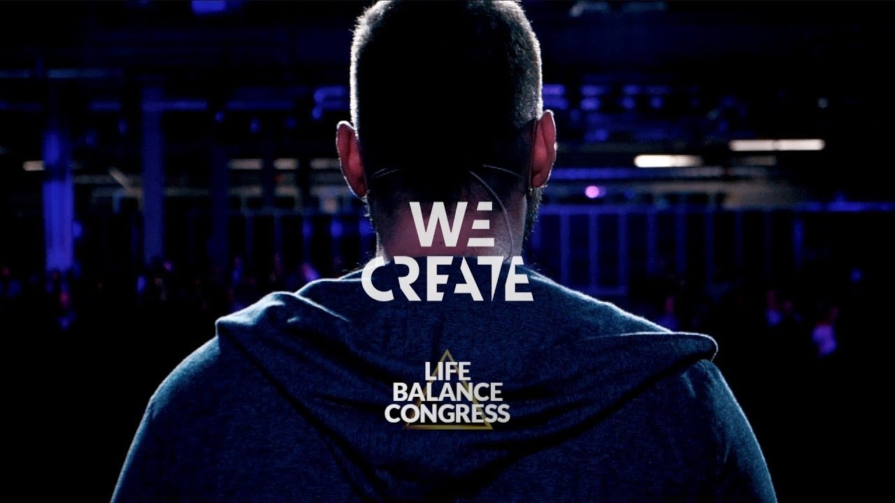 Widok zza pleców osoby z mikrofonem nagłownym, skierowanej w stronę publiczności na konferencji Life Balance Congress, z napisem 'We Create' na ubraniu.