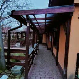 CarsVendors Szymon Nowak - Zadaszony drewniany ganek przylegający do pomarańczowej ściany budynku, wyłożony kostką brukową, z widocznym fragmentem ogrodu i drzewa bez liści w tle.