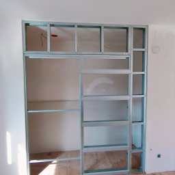 CarsVendors Szymon Nowak - Metalowa konstrukcja ścianki działowej z profili, widoczne kable elektryczne, surowa ściana i panele podłogowe w jasnym pomieszczeniu w trakcie prac wykończeniowych.