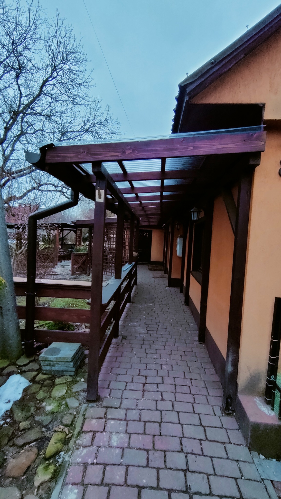 Zadaszony drewniany ganek przylegający do pomarańczowej ściany budynku, wyłożony kostką brukową, z widocznym fragmentem ogrodu i drzewa bez liści w tle.