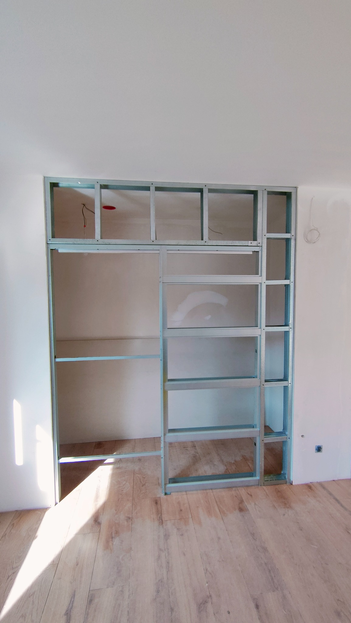 Metalowa konstrukcja ścianki działowej z profili, widoczne kable elektryczne, surowa ściana i panele podłogowe w jasnym pomieszczeniu w trakcie prac wykończeniowych.