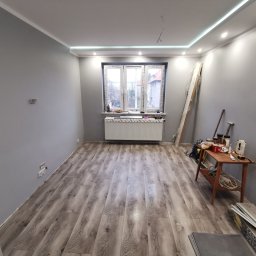 Klementowski Remonty - Pokój po remoncie z szarą podłogą imitującą drewno, szarymi ścianami, białym sufitem z oświetleniem LED, grzejnikiem pod oknem i stolikiem z narzędziami.