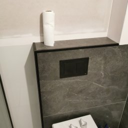 Klementowski Remonty - Fragment toalety podtynkowej z szarą płytką imitującą kamień, czarnym przyciskiem spłukującym Tece i rolką papieru toaletowego na półce. Widoczne białe silikony na sedesie.