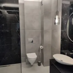 Nowoczesna łazienka z szarymi płytkami, czarnym marmurem pod prysznicem, wiszącą toaletą i owalną umywalką na czarnej szafce, oświetlona wiszącą lampą z żarówką Edison