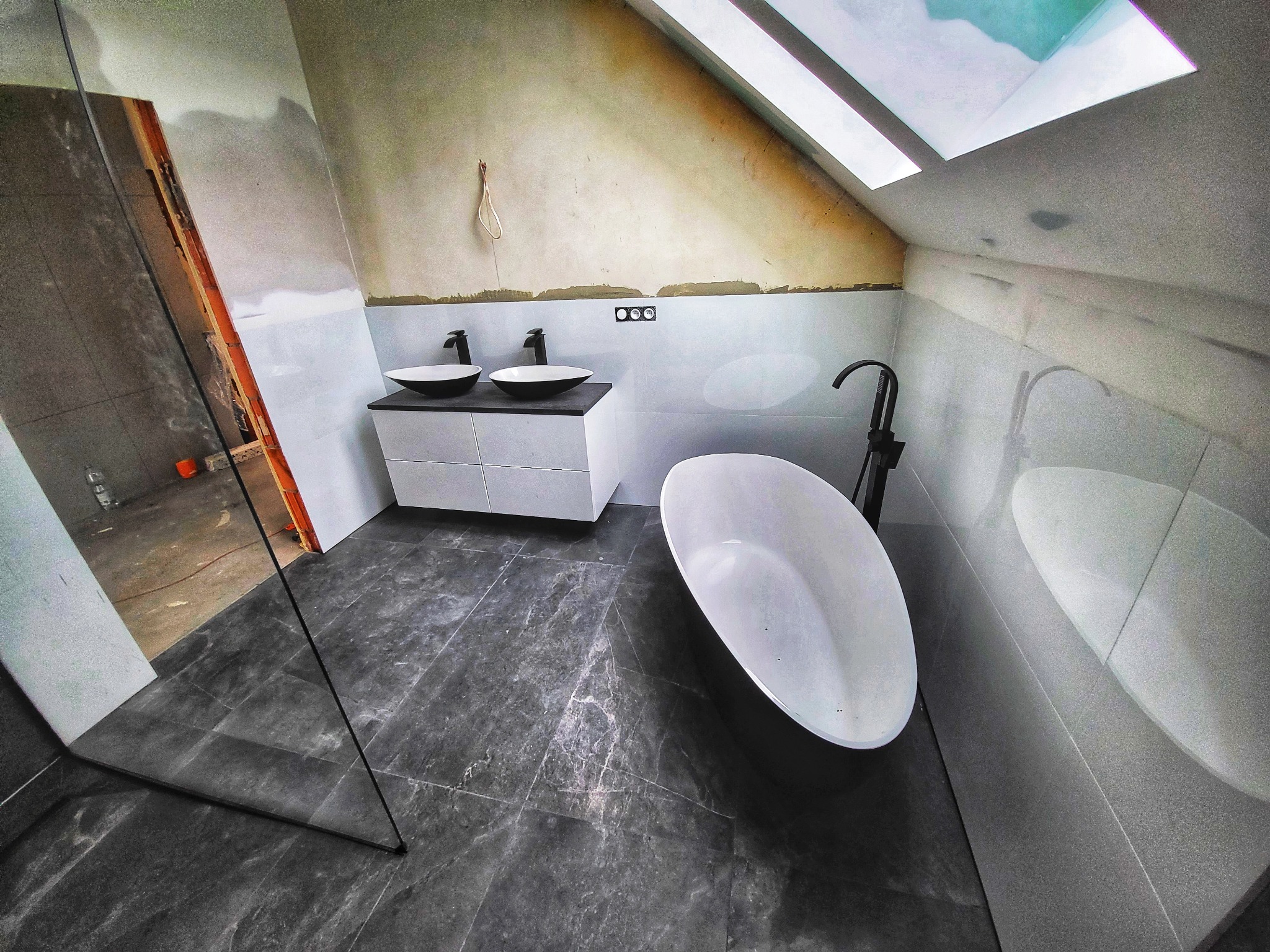 Niedokończona łazienka na poddaszu z czarnymi bateriami, białą wanną wolnostojącą i podwójną umywalką na białej szafce, szare płytki podłogowe i ściany częściowo wykończone białymi płytkami...