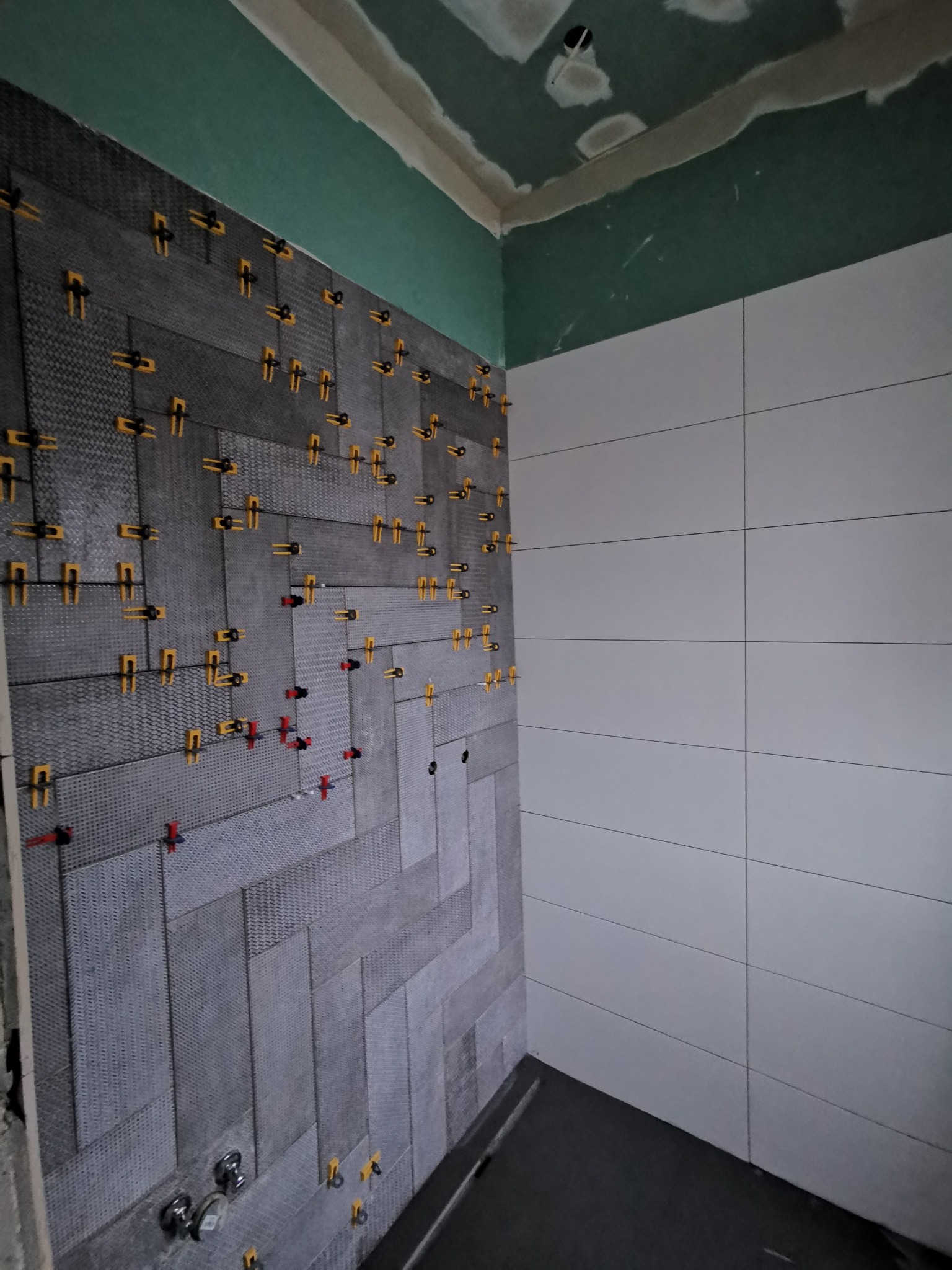 Układanie szarej, wzorzystej glazury w jodełkę z użyciem klipsów poziomujących na ścianie łazienki, widoczna ściana z jasnymi, poziomymi płytkami oraz surowy sufit w kolorze zielonym.