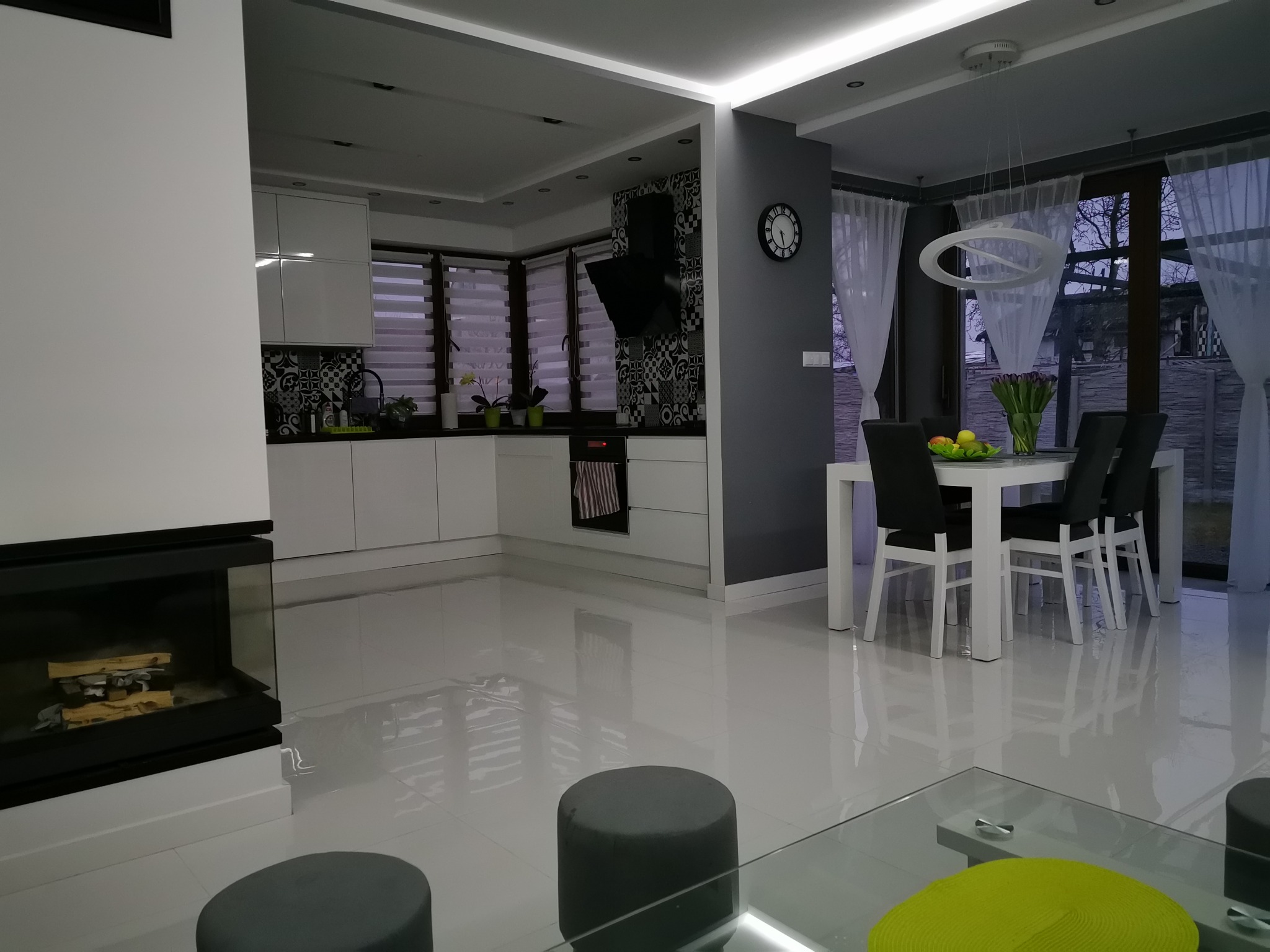 Nowoczesne wnętrze: biała kuchnia z czarnymi akcentami, kominek z palącym się drewnem, stół jadalny z bukietem tulipanów, szare pufy i błyszcząca podłoga z odbiciami światła.