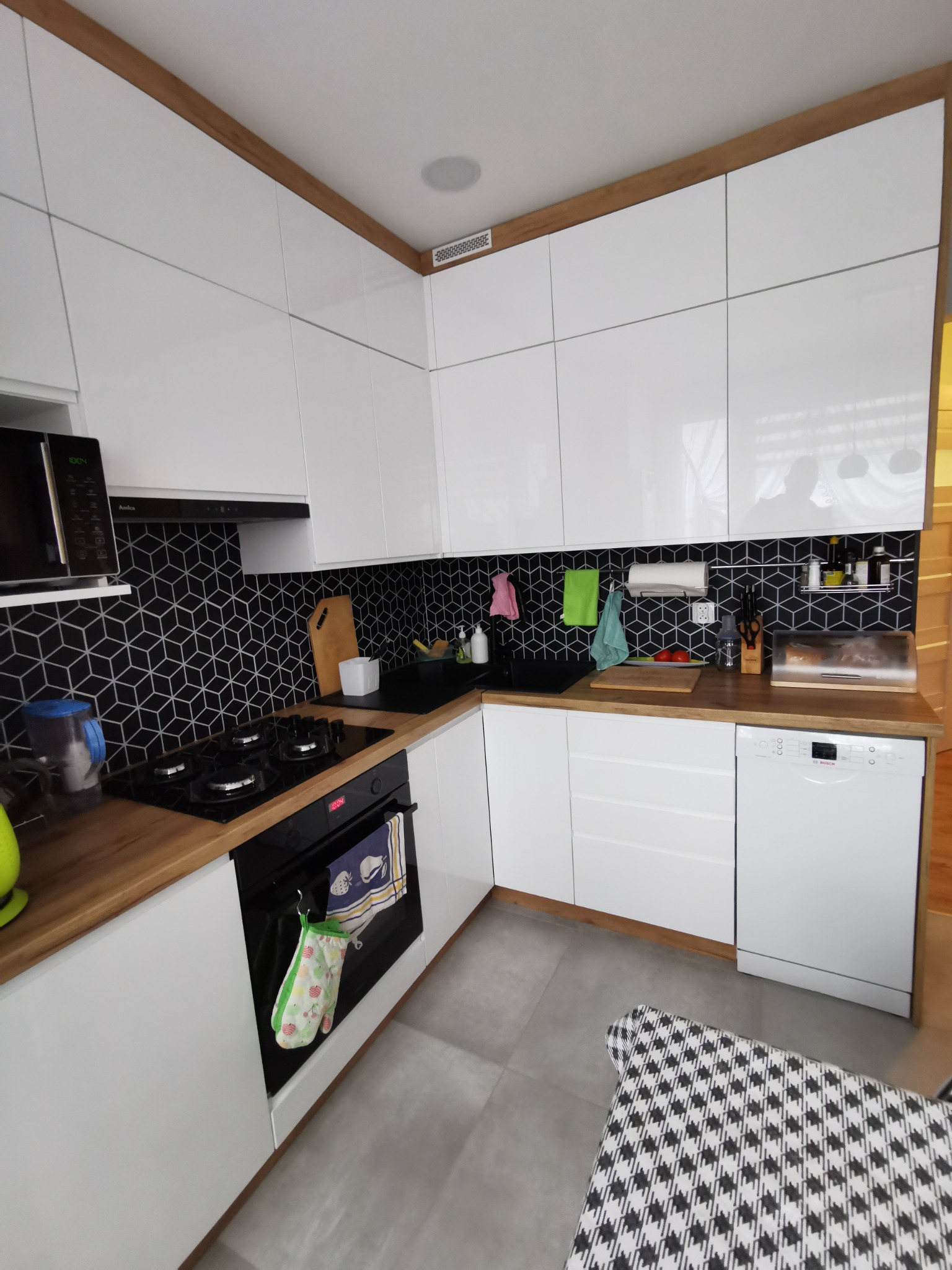 Nowoczesna, jasna kuchnia z białymi szafkami i drewnianymi blatami w Dzierżoniowie. Uwagę przyciągają czarne, geometryczne płytki na ścianie nad blatem. Na blacie widoczne akcesoria kuchenne...