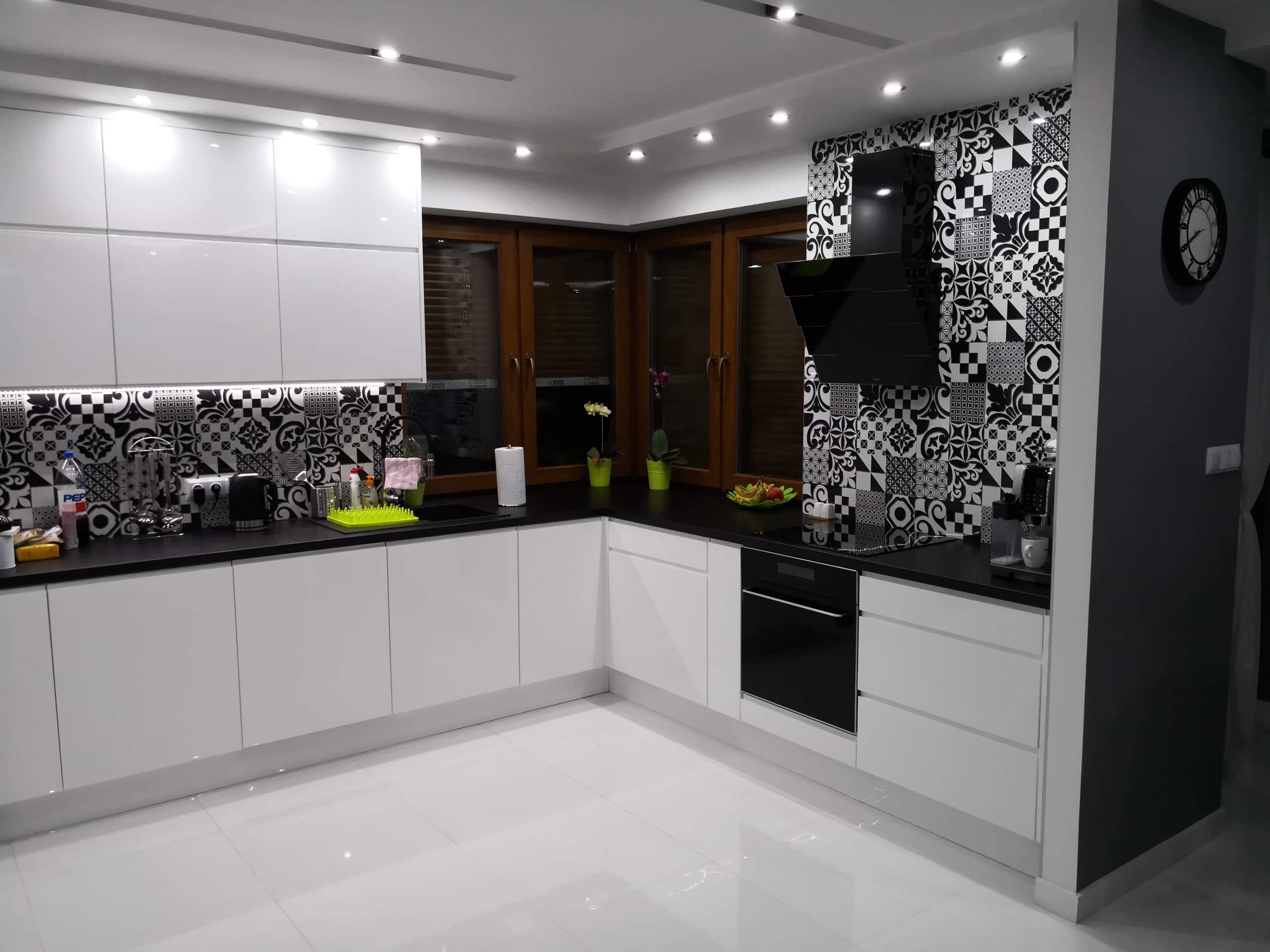 Nowoczesna, jasna kuchnia z białymi szafkami, czarnym blatem, geometrycznymi czarno-białymi płytkami na ścianie, drewnianymi oknami i czarnym okapem.