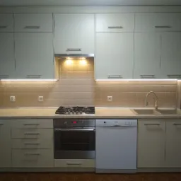 Jasna kuchnia z beżowymi płytkami nad blatem, oświetleniem LED pod szafkami wiszącymi i brązową podłogą, z widoczną zmywarką i piekarnikiem.