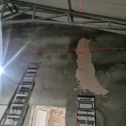 Surowy mur z zamontowaną skrzynką elektryczną, wyznaczona laserem pionowa i pozioma linia, drabina oparta o ścianę, widoczne elementy konstrukcji dachu.