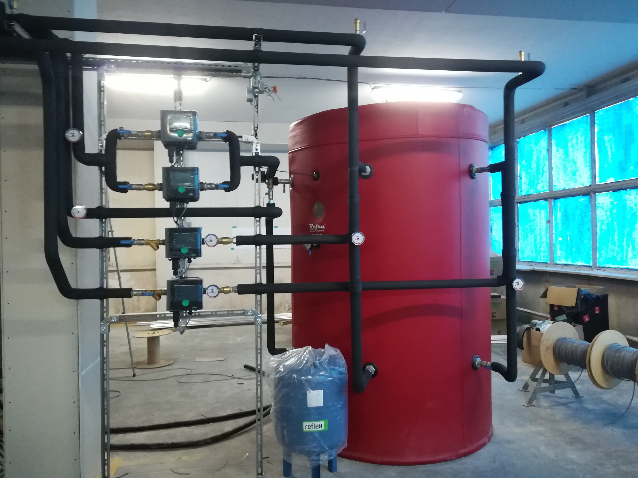 Czerwony zbiornik akumulacyjny ciepła z czarnymi rurami, manometrami i elektronicznymi regulatorami, w tle okno z niebieską folią, na podłodze mniejszy niebieski zbiornik w folii i szpule...