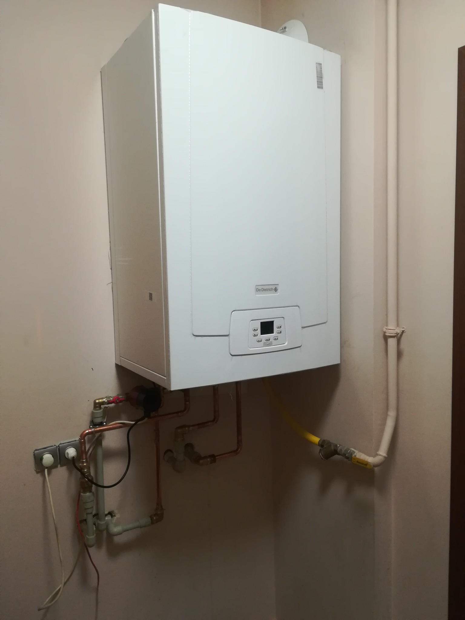 Biały, wiszący piec gazowy marki De Dietrich zamontowany na ścianie, widoczne podłączenia hydrauliczne z rur miedzianych i plastikowych, oraz przyłącze gazowe z żółtym przewodem.