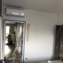 KKBuilding - Pokój w trakcie przygotowań do malowania: ściany zabezpieczone taśmą malarską i folią, widoczny klimatyzator, ślady po szpachlowaniu na ścianie.