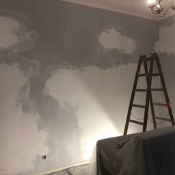 KKBuilding - Ściana w trakcie malowania, z widocznymi plamami szarej farby i drewnianą drabiną opartą o ścianę, zabezpieczone meble pod ścianą, widoczny żyrandol na suficie.