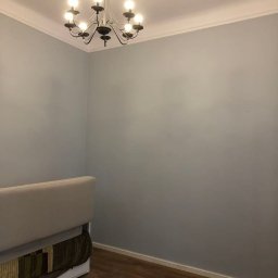 KKBuilding - Pomalowane na szaro pomieszczenie z drewnianą podłogą, widoczny fragment sofy i ozdobny żyrandol z żarówkami w stylu świec.